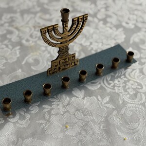 May include: A brass menorah with nine candle holders, set on a rectangular blue base. The central holder is taller than the others. The menorah is placed on a white floral patterned surface.