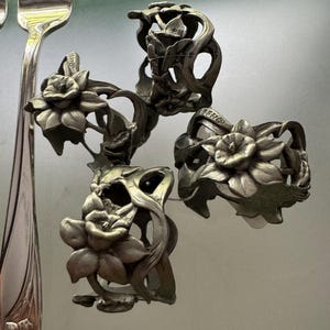 Pu&ograve; includere: Un set di tre portatovaglioli in metallo color argento con un design a rilievo di fiori di narciso con steli e foglie intricati, accompagnato da una forchetta in metallo color argento abbinata con un motivo di narciso sul manico.