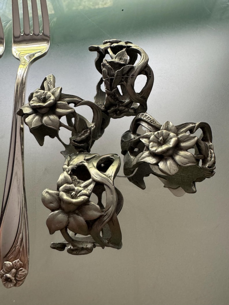 Pu&ograve; includere: Un set di tre portatovaglioli in metallo color argento con un design a rilievo di fiori di narciso con steli e foglie intricati, accompagnato da una forchetta in metallo color argento abbinata con un motivo di narciso sul manico.