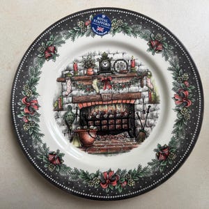 以下が含まれることがあります： ロイヤルスタッフォードの装飾用プレート。クリスマスの暖炉のシーンが描かれています。プレートの黒い縁には花とベリーのデザイン。中央には、靴下、時計、火のある暖炉の詳細なイラストが描かれています。