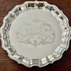 May include: A round silver-toned metal serving tray with a scalloped edge and a reflective surface. The center of the tray features an intricate engraved design with the text "Happy Anniversary" written in a decorative cursive script.