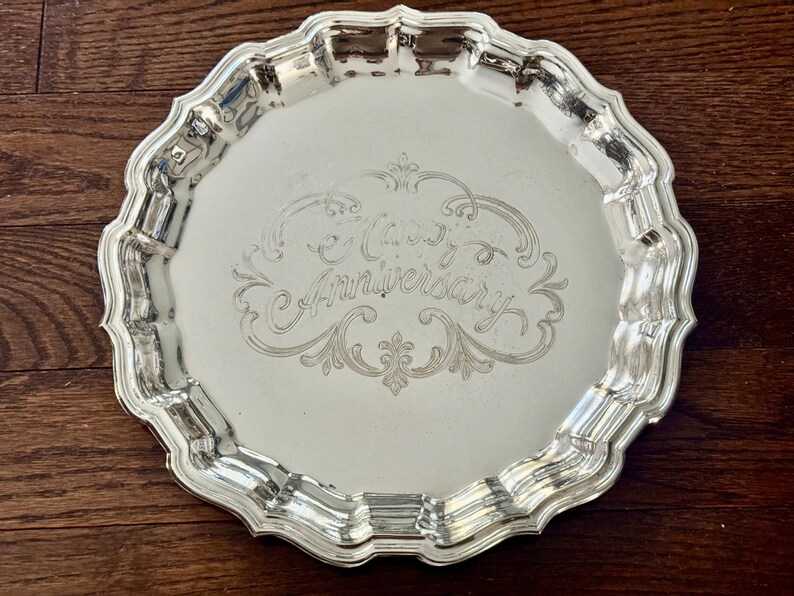 May include: A round silver-toned metal serving tray with a scalloped edge and a reflective surface. The center of the tray features an intricate engraved design with the text "Happy Anniversary" written in a decorative cursive script.