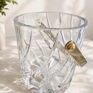 May include: A clear glass ice bucket featuring a faceted, geometric cut pattern across its exterior surface. The bucket includes a metallic handle attached to the sides with gold-toned circular hardware, creating a reflective and textured appearance.