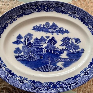 K&ouml;nnte beinhalten: Eine wei&szlig;e ovale Keramik-Servierplatte mit einem blauen und wei&szlig;en Weidenmuster. Das Muster zeigt eine Br&uuml;cke, B&auml;ume und einen Pagodentempel.