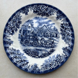 May include: A round ceramic plate with a scalloped edge featuring a dark blue and white transferware pattern. The center depicts a horse-drawn stagecoach in front of stone buildings, while the border displays intricate floral and scroll motifs.