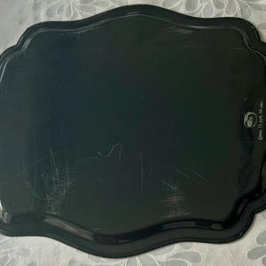 May include: A black, scalloped-edge tray with a glossy finish. The tray has a slightly irregular shape and appears to be made of metal or a similar material. There are some scratches on the surface. The tray has a decorative design.