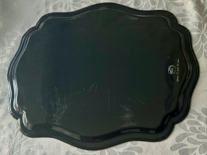 May include: A black, scalloped-edge tray with a glossy finish. The tray has a slightly irregular shape and appears to be made of metal or a similar material. There are some scratches on the surface. The tray has a decorative design.