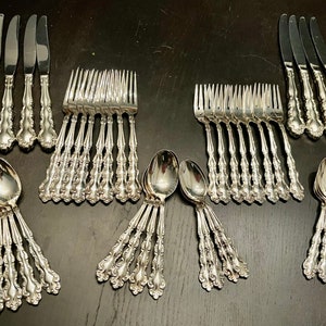 K&ouml;nnte beinhalten: Ein Set aus silbernem Besteck, bestehend aus Gabeln, Messern und L&ouml;ffeln, mit verzierten Designs. Das Besteck ist in Reihen auf einer schwarzen Oberfl&auml;che angeordnet.