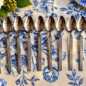 May include: A set of ten silver-toned metal spoons arranged in a row. Each spoon features a long, rectangular handle with a detailed, embossed geometric and floral pattern running down the centre. The bowls of the spoons are smooth and oval.