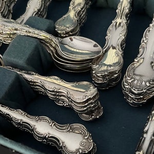 May include: Close-up of a collection of ornate silver spoons and serving spoons. The silverware has a detailed, floral design on the handles and is arranged on a dark blue surface. The spoons are stacked in neat rows.