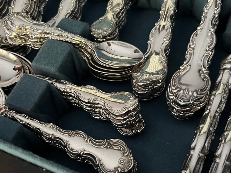 May include: Close-up of a collection of ornate silver spoons and serving spoons. The silverware has a detailed, floral design on the handles and is arranged on a dark blue surface. The spoons are stacked in neat rows.