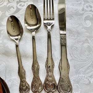 May include: A set of vintage silverware, including two spoons, a fork, and a knife. The silverware is silver-colored with ornate floral detailing on the handles. The pieces are arranged on a white, patterned tablecloth.