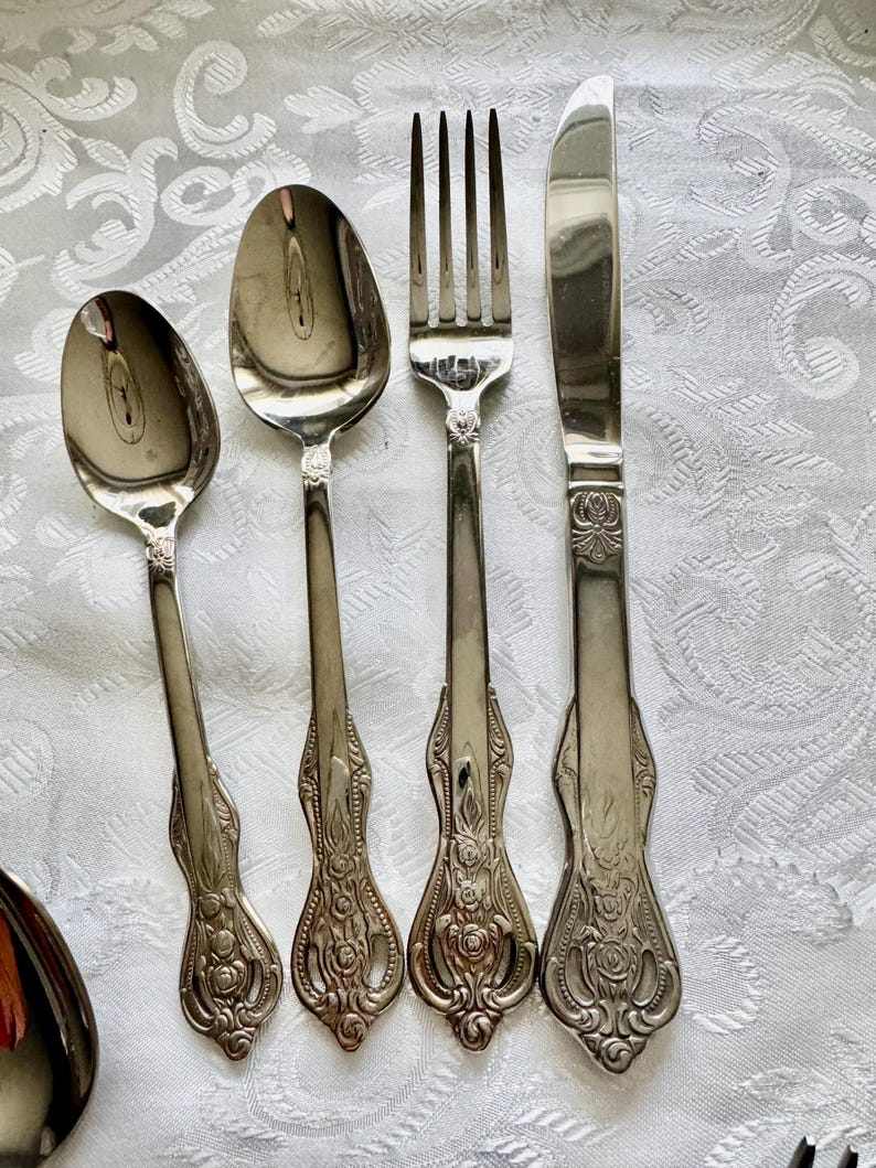 May include: A set of vintage silverware, including two spoons, a fork, and a knife. The silverware is silver-colored with ornate floral detailing on the handles. The pieces are arranged on a white, patterned tablecloth.