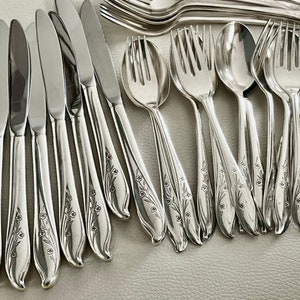 May include: A collection of shiny silver flatware, including knives, forks, and spoons. The silverware features a floral design on the handles. The utensils are arranged on a light-colored surface, suggesting a formal dining setting.