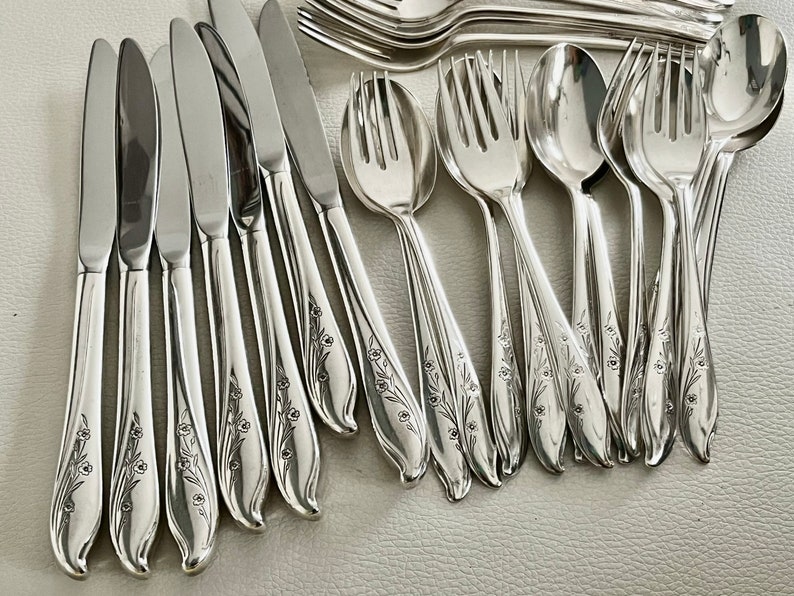 May include: A collection of shiny silver flatware, including knives, forks, and spoons. The silverware features a floral design on the handles. The utensils are arranged on a light-colored surface, suggesting a formal dining setting.