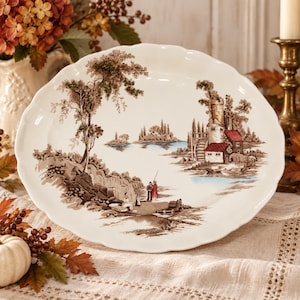 May include: An oval ceramic platter with a scalloped edge featuring a brown and white transferware landscape scene of a lake, stone buildings with red roofs, and figures standing on a rocky shore near a tall tree.