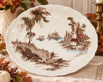 May include: An oval ceramic platter with a scalloped edge featuring a brown and white transferware landscape scene of a lake, stone buildings with red roofs, and figures standing on a rocky shore near a tall tree.