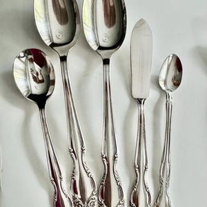 May include: A collection of polished silver flatware, including spoons and a butter knife, arranged on a white surface. The spoons vary in size, with ornate floral detailing on the handles. The butter knife has a rounded blade. The silverware reflects the surrounding environment.
