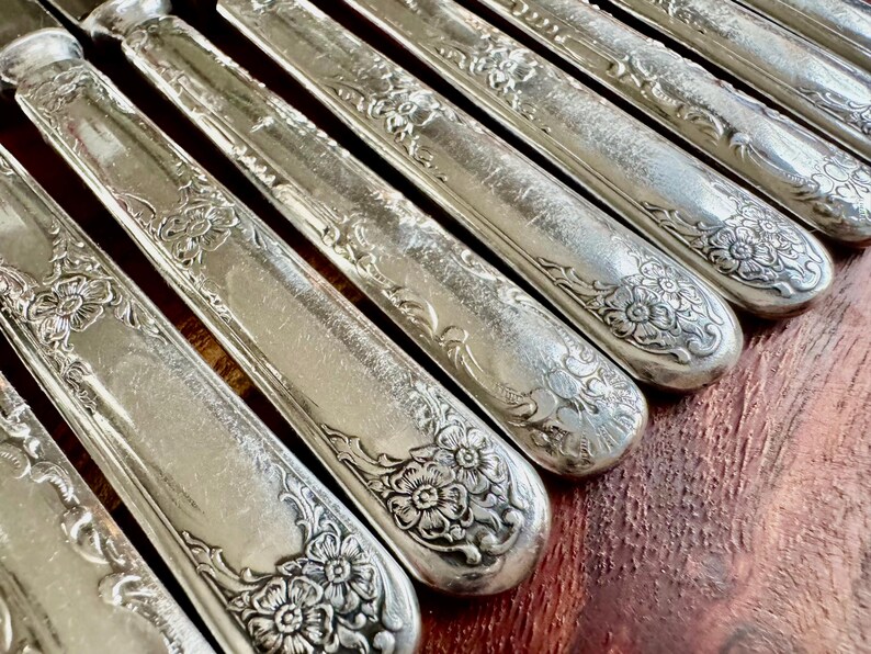 May include: A set of silver-plated dinner knives with ornate floral designs on the handles. The knives are arranged in a row, with the handles pointing to the right.
