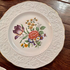 May include: A round white ceramic plate featuring a central floral bouquet with purple, red, yellow, and blue flowers and green leaves. The wide rim has an embossed lattice and scroll pattern with a thin gold-painted edge along the scalloped border.