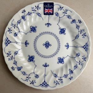 Puede incluir: Un plato de cer&aacute;mica blanca con borde festoneado, con un patr&oacute;n floral azul y un motivo circular central. Una etiqueta rectangular en la parte superior muestra el texto Queen's by Churchill, una bandera brit&aacute;nica y Made in England.