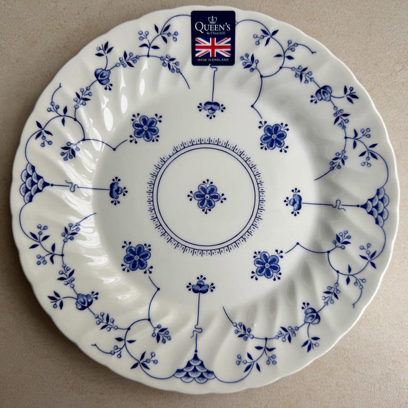 Puede incluir: Un plato de cer&aacute;mica blanca con borde festoneado, con un patr&oacute;n floral azul y un motivo circular central. Una etiqueta rectangular en la parte superior muestra el texto Queen's by Churchill, una bandera brit&aacute;nica y Made in England.