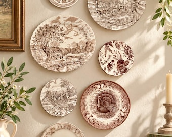 May include: A collection of seven decorative ceramic plates mounted on a wall, featuring brown and white transferware patterns including rural landscapes, floral motifs, a turkey, and a rabbit, all with intricate scalloped or smooth edges.