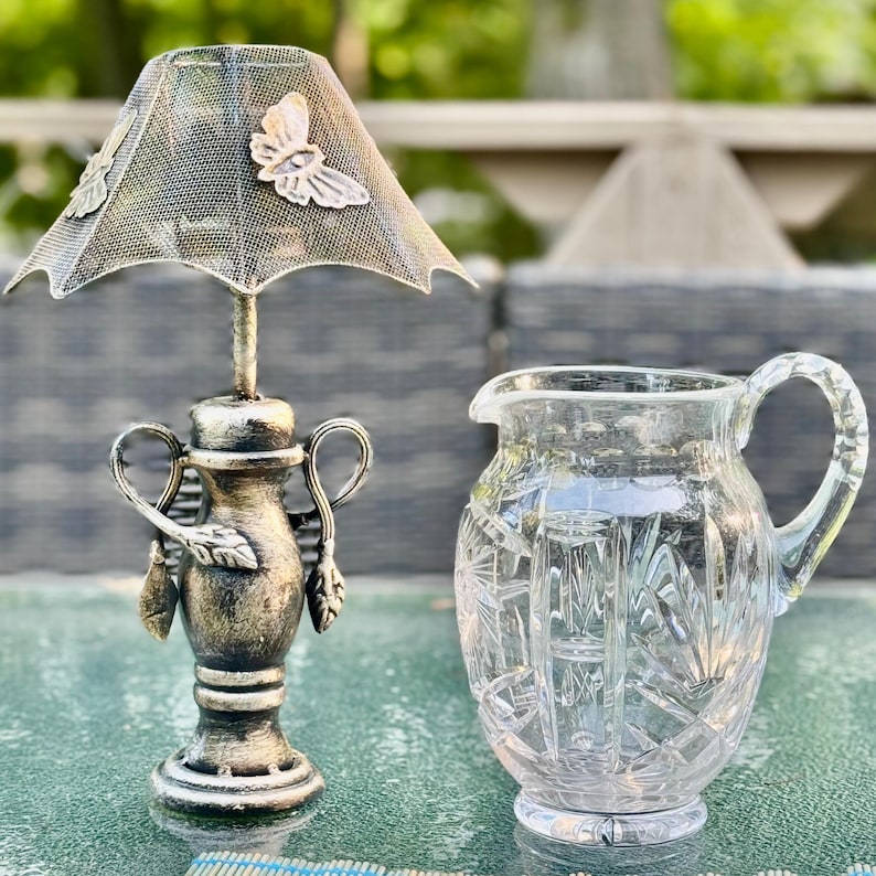 Puede incluir: Un portavelas decorativo de metal con una pantalla en forma de mariposa y una jarra de vidrio transparente. El portavelas tiene un acabado bronce con detalles de hojas. La jarra tiene un dise&ntilde;o de cristal tallado.