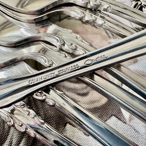 May include: Close-up of a collection of Community Stainless silverware. The image highlights the shiny, reflective surfaces of the forks and spoons. The text "Community Stainless Oneida" is visible on one of the utensils. The silverware has a decorative, ornate design.