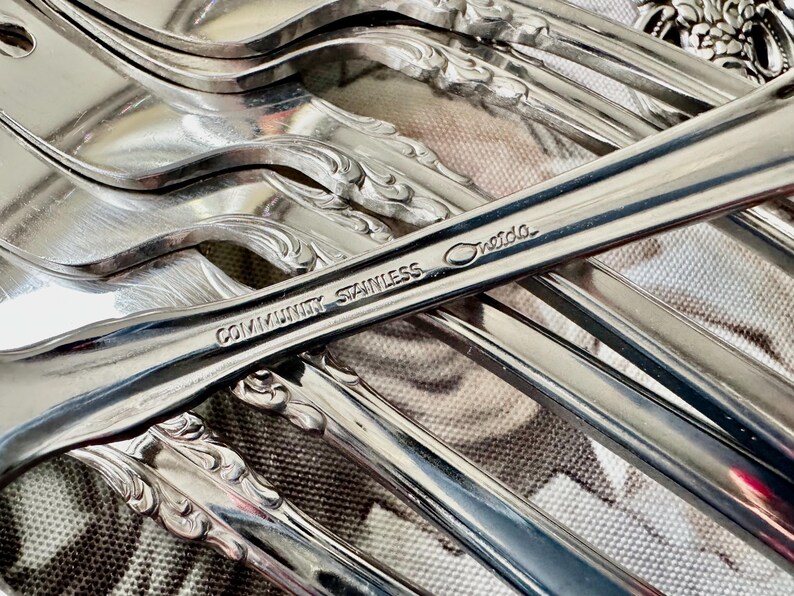 May include: Close-up of a collection of Community Stainless silverware. The image highlights the shiny, reflective surfaces of the forks and spoons. The text "Community Stainless Oneida" is visible on one of the utensils. The silverware has a decorative, ornate design.