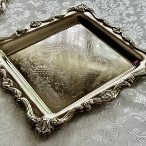 Puede incluir: Una bandeja cuadrada de metal plateado con un borde elevado y ornamentado que presenta intrincados motivos de volutas y formas de concha. La superficie central es lisa y reflectante, con un sutil patr&oacute;n grabado en los bordes internos.
