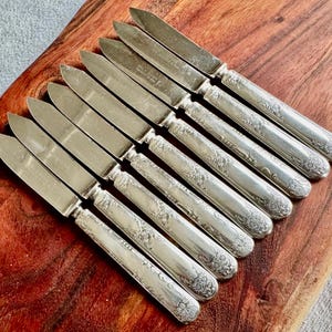 May include: A set of ten silver butter knives with ornate handles. The knives are arranged on a wooden surface.