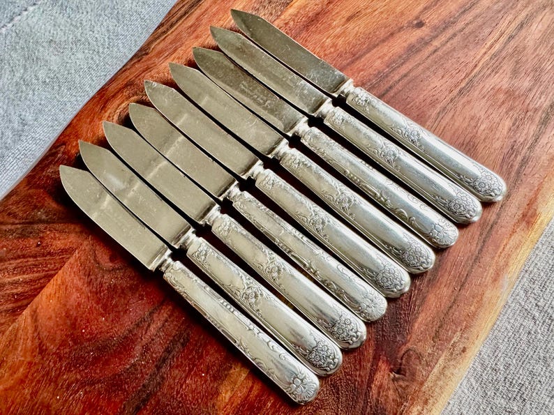 May include: A set of ten silver butter knives with ornate handles. The knives are arranged on a wooden surface.