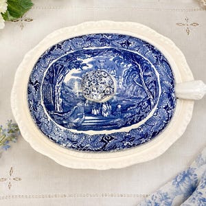 Op de afbeelding: Een ovaal keramisch deksel voor een serveerschaal met een cr&egrave;mekleurige geschulpte rand en een centrale blauw-witte transferware-sc&egrave;ne met een kasteel, bomen en figuren. Het deksel heeft een bloemmotief knop en een wit handvat aan de zijkant.