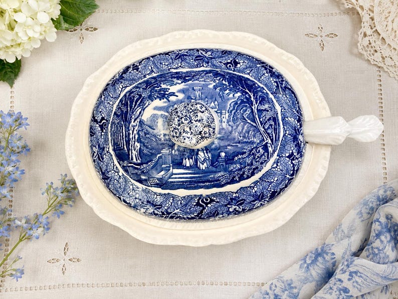 Op de afbeelding: Een ovaal keramisch deksel voor een serveerschaal met een cr&egrave;mekleurige geschulpte rand en een centrale blauw-witte transferware-sc&egrave;ne met een kasteel, bomen en figuren. Het deksel heeft een bloemmotief knop en een wit handvat aan de zijkant.