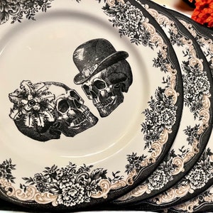 Puede incluir: Un juego de platos de cer&aacute;mica color crema con una ilustraci&oacute;n en blanco y negro de dos calaveras, una con bomb&iacute;n y otra adornada con un tocado floral. Los bordes est&aacute;n decorados con un detallado patr&oacute;n floral en negro y beige.