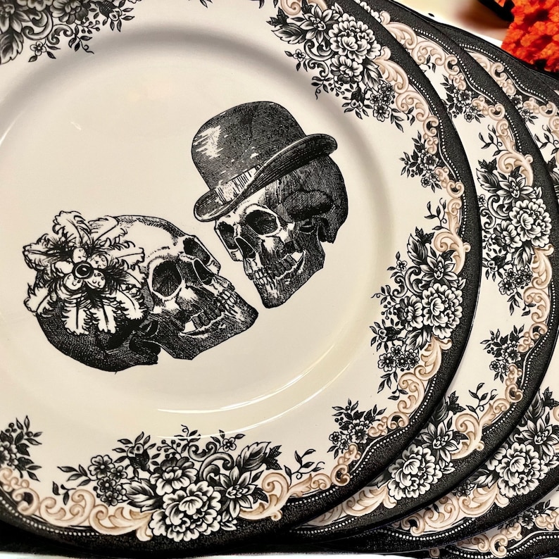 Puede incluir: Un juego de platos de cer&aacute;mica color crema con una ilustraci&oacute;n en blanco y negro de dos calaveras, una con bomb&iacute;n y otra adornada con un tocado floral. Los bordes est&aacute;n decorados con un detallado patr&oacute;n floral en negro y beige.