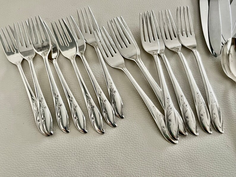 May include: A collection of silver forks with floral detailing on the handles. The forks are arranged on a light-colored surface, showcasing their polished finish. The tines are evenly spaced, and the design suggests a formal dining setting.