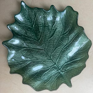 Puede incluir: Un cuenco de cristal verde oscuro con forma de hoja grande y superficie texturizada que imita las venas naturales y los contornos org&aacute;nicos. El cuenco presenta un acabado brillante y bordes irregulares festoneados que siguen la silueta de una hoja.