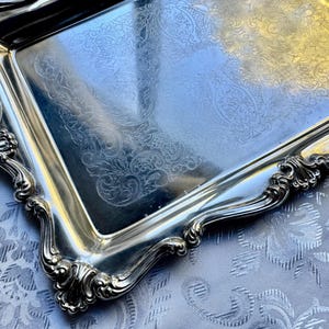 Puede incluir: Una bandeja de servicio rectangular de tono plateado con una superficie pulida y reflectante que presenta un intrincado patr&oacute;n grabado de flores y volutas. La bandeja tiene un borde elevado y festoneado con motivos en relieve.