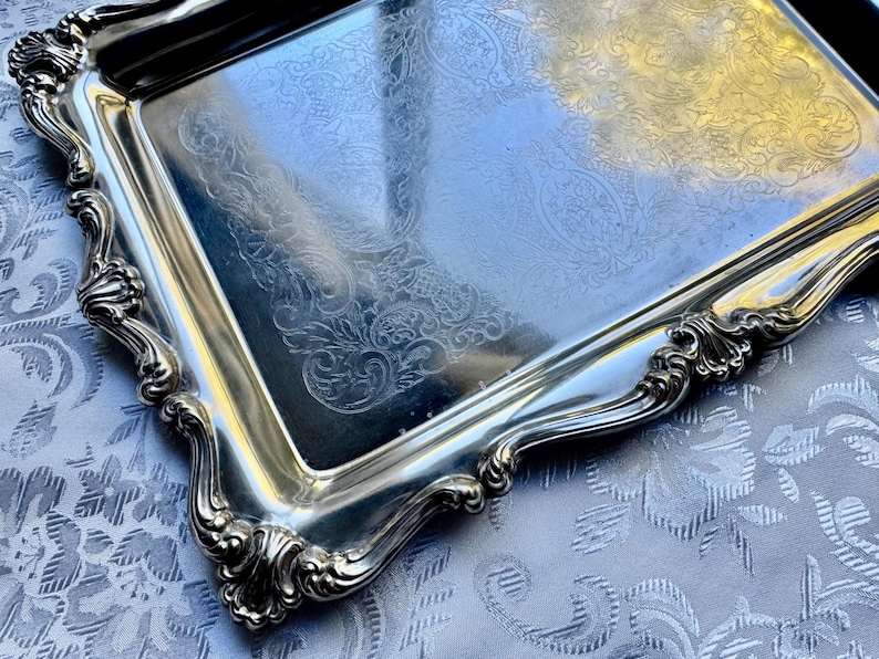Puede incluir: Una bandeja de servicio rectangular de tono plateado con una superficie pulida y reflectante que presenta un intrincado patr&oacute;n grabado de flores y volutas. La bandeja tiene un borde elevado y festoneado con motivos en relieve.