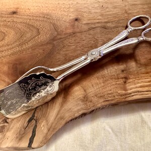 May include: Silver serving tongs with an ornate, engraved design. The tongs have a scalloped edge and are resting on a wooden surface.