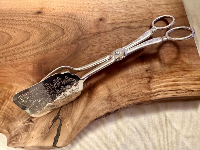 May include: Silver serving tongs with an ornate, engraved design. The tongs have a scalloped edge and are resting on a wooden surface.