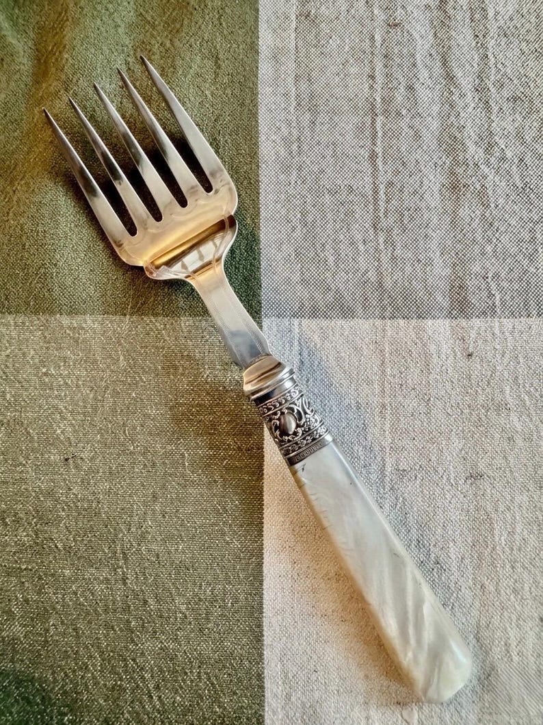 May include: Silver serving fork with six tines and a decorative handle. The handle is white with a pearlescent sheen, and the metal collar features an ornate design. The fork is set against a green and white checkered background.
