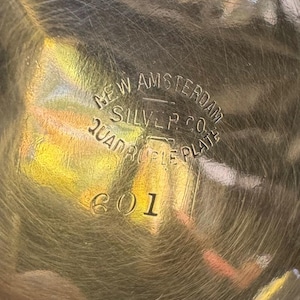 May include: A metallic surface with a circular engraved hallmark reading NEW AMSTERDAM SILVER CO. QUADRUPLE PLATE and the number 601. The surface is reflective with visible fine scratches and a golden hue, showing clear stamped lettering.