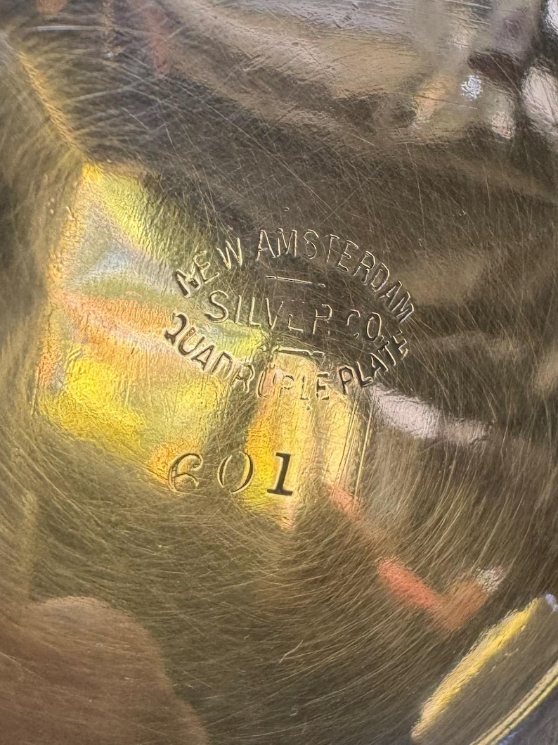 May include: A metallic surface with a circular engraved hallmark reading NEW AMSTERDAM SILVER CO. QUADRUPLE PLATE and the number 601. The surface is reflective with visible fine scratches and a golden hue, showing clear stamped lettering.