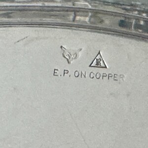 May include: A silver-toned metal surface featuring stamped hallmarks including a fox head icon and a letter R inside a triangle. Below these symbols, the text E.P. ON COPPER is clearly engraved into the metallic material of the object.