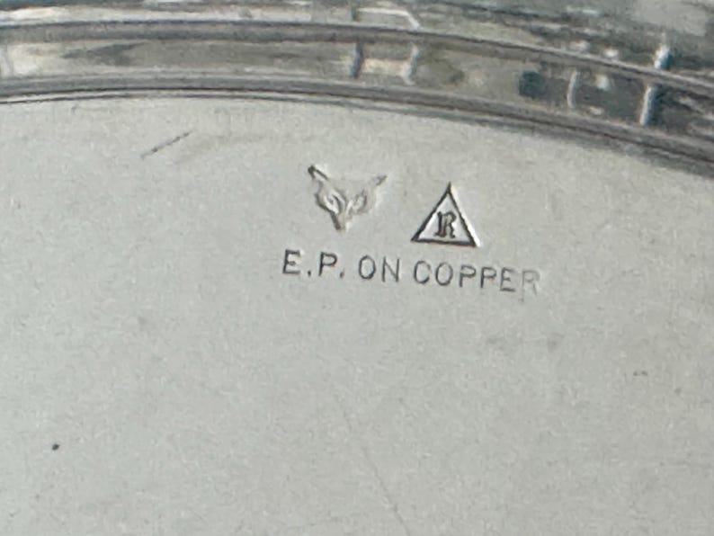 May include: A silver-toned metal surface featuring stamped hallmarks including a fox head icon and a letter R inside a triangle. Below these symbols, the text E.P. ON COPPER is clearly engraved into the metallic material of the object.