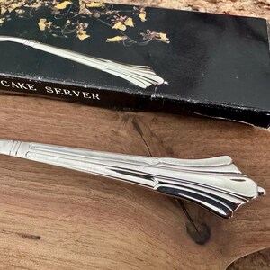 K&ouml;nnte beinhalten: Ein silberfarbener Tortenheber aus Metall mit einem dekorativen, geriffelten Griff. Der Tortenheber liegt vor seiner originalen, schwarzen, rechteckigen Verpackung, die mit einem Blumenmuster und dem Text PLATED CAKE SERVER bedruckt ist.