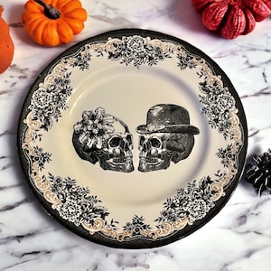 Puede incluir: Un plato redondo de cer&aacute;mica con un centro de color crema que presenta dos calaveras negras frente a frente, una con un tocado floral y la otra con un bomb&iacute;n. El borde est&aacute; decorado con un patr&oacute;n floral negro y crema y volutas.